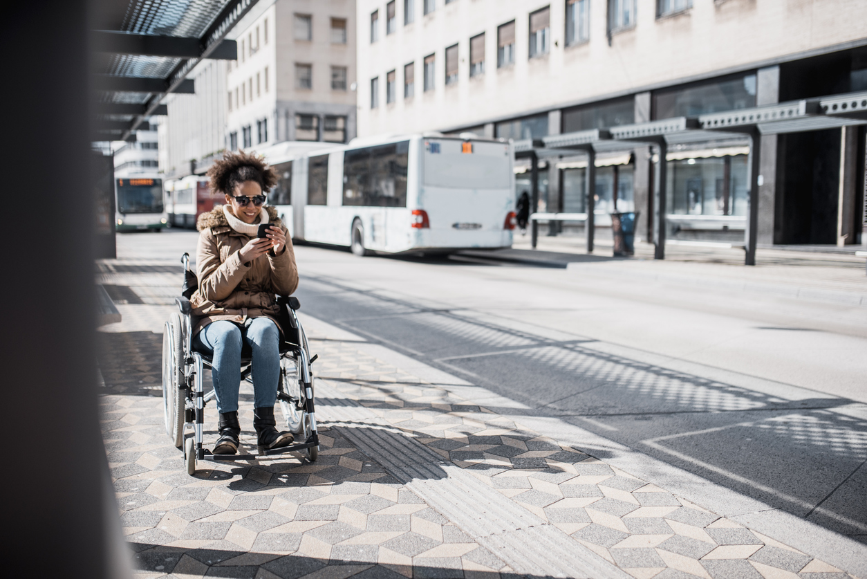 A woman using a smartphone in a wheelchair at a bus stop. A bus is in the background