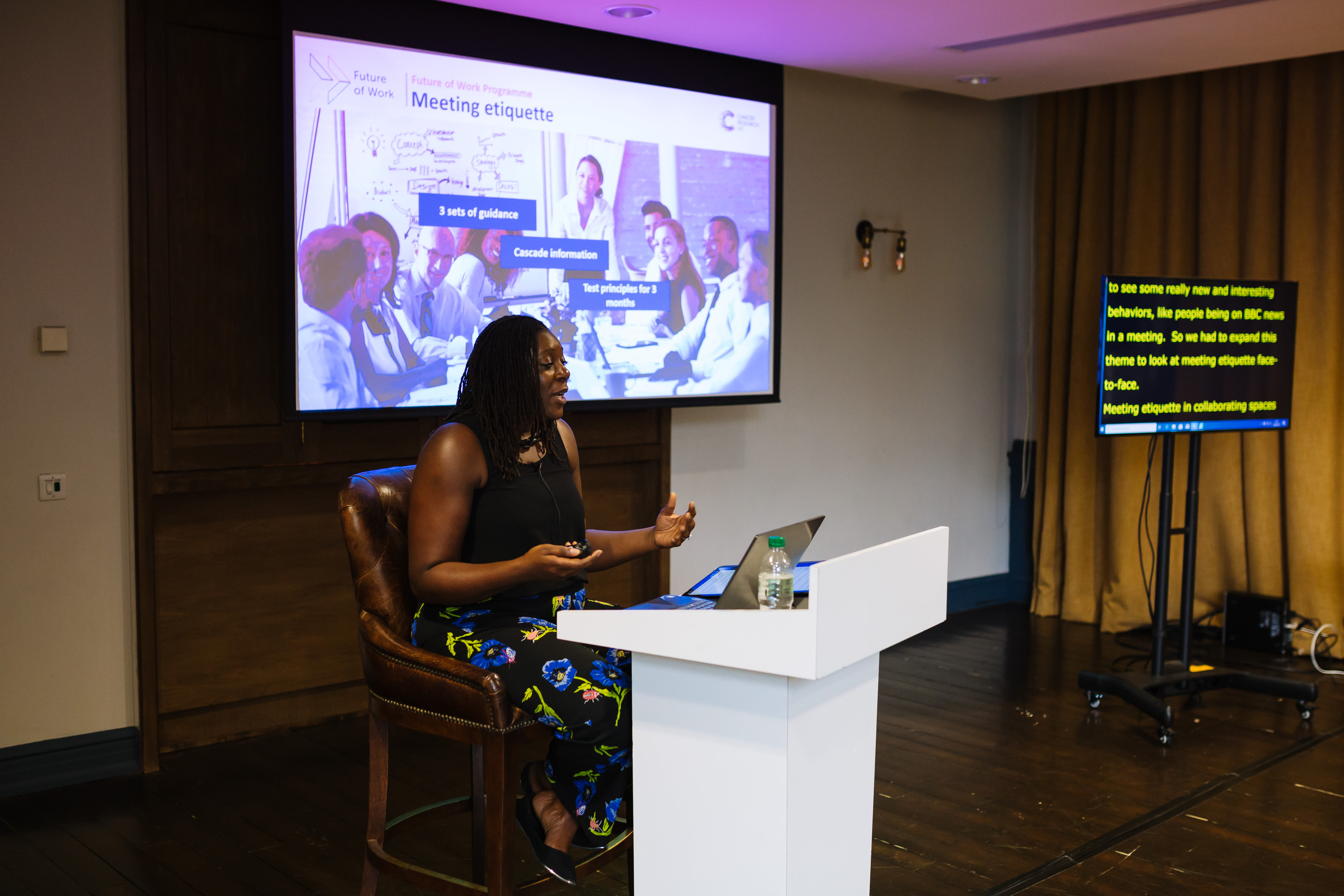 Rachel Xavier. A black woman with mid length hair sat on a stool behind a podium. She is presenting with a large presenting screen displaying "Meeting Etiquette" behind her, and there's a subtitles screen to the right. 
