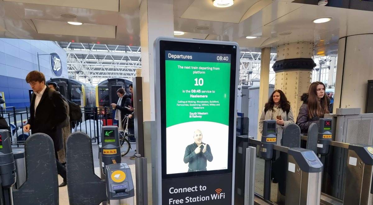 A screen shows an AI sign language tool in operation at a ticket gate, the AI generated person on screen is signing with corresponding text above