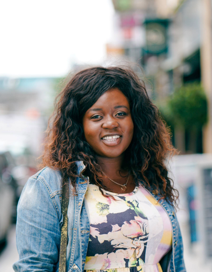 Headshot of Camp Digital speaker Candi Williams. Candi is standing outdoors on a city street, wearing a light blue denim jacket over a colorful floral-patterned dress. The background shows greenery and buildings, suggesting a lively street scene.