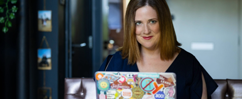 Sharon O'Dea sits at a desk. The back of her laptop is visible, and covered in stickers. She has short brown hair and blue eyes.