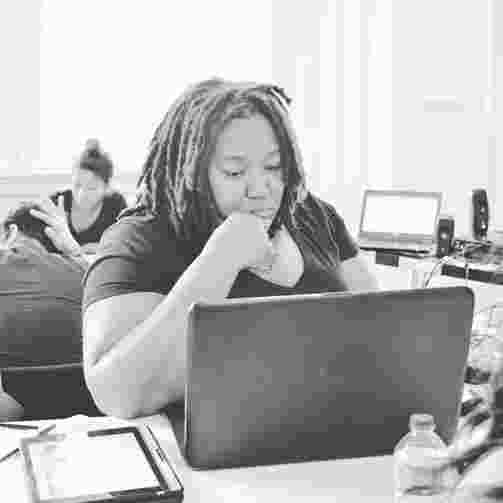 A black and white photo of a black woman with dreadlocks, looking glum and staring at a laptop. 