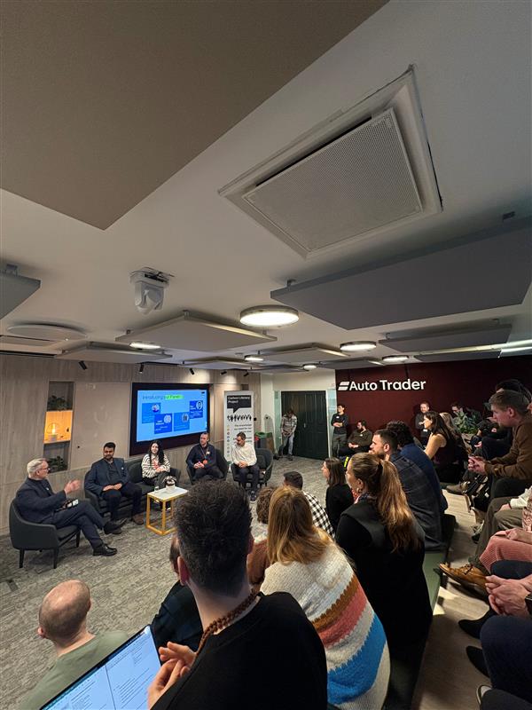 A seated crowd of people look at a panel of four people sitting in chairs. Behind them the Auto Trader logo is visible. 