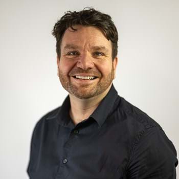 Hugh is standing against a plain white background and smiling. He has short dark hair and a beard, wearing a dark button-up shirt