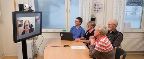 Three older white people with a white nurse sat around a table looking at a large television on a mobile stand. They are on a video conference and a white lady's face is displayed on the tv screen.