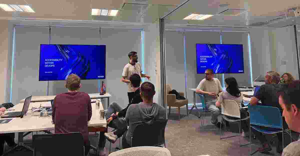 Danny Lancaster standing in front of a room of seated people. He is speaking, and on two screens is a slide reading "Accessibility within DevOps."