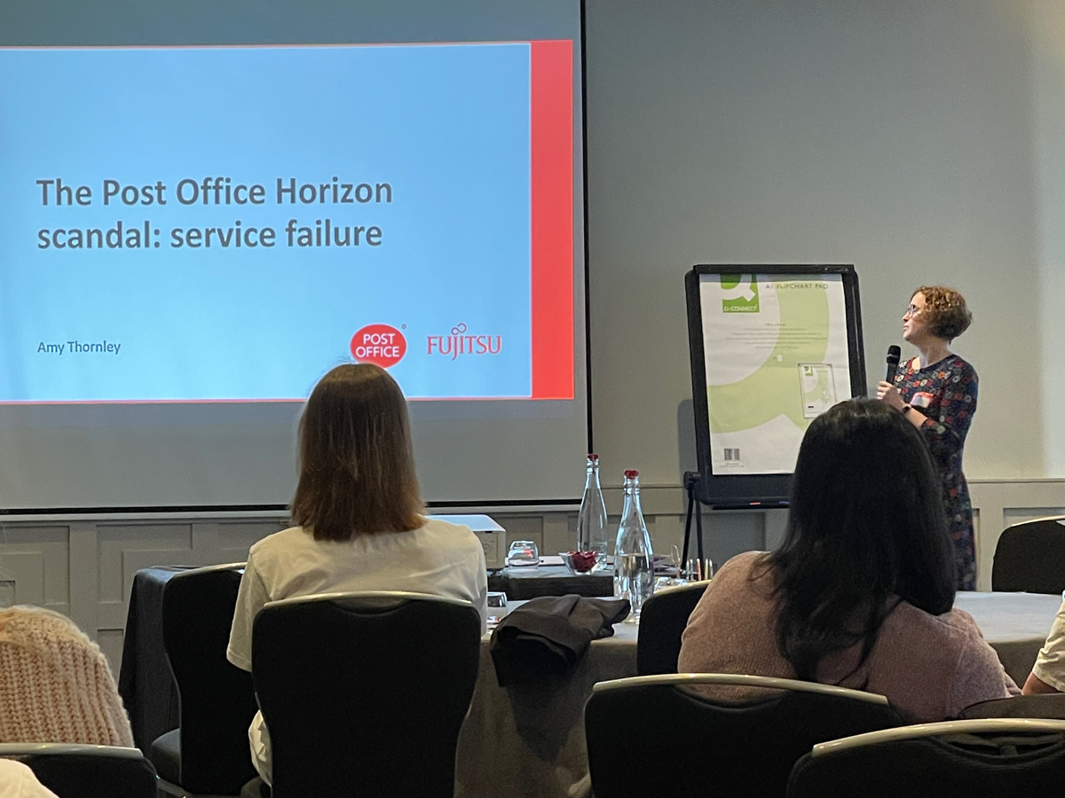 Amy stands delivering a presentation to the team. Onscreen is a title slide: The Post Office Horizon Scandal: Service failure
