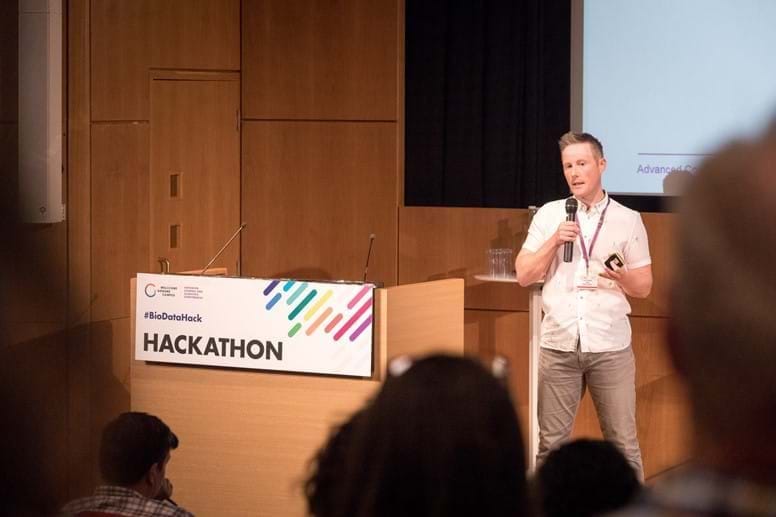 Francis Rowland, a ginger haired man with a white shirt and grey chinos, presenting at the BioData Hackathon.