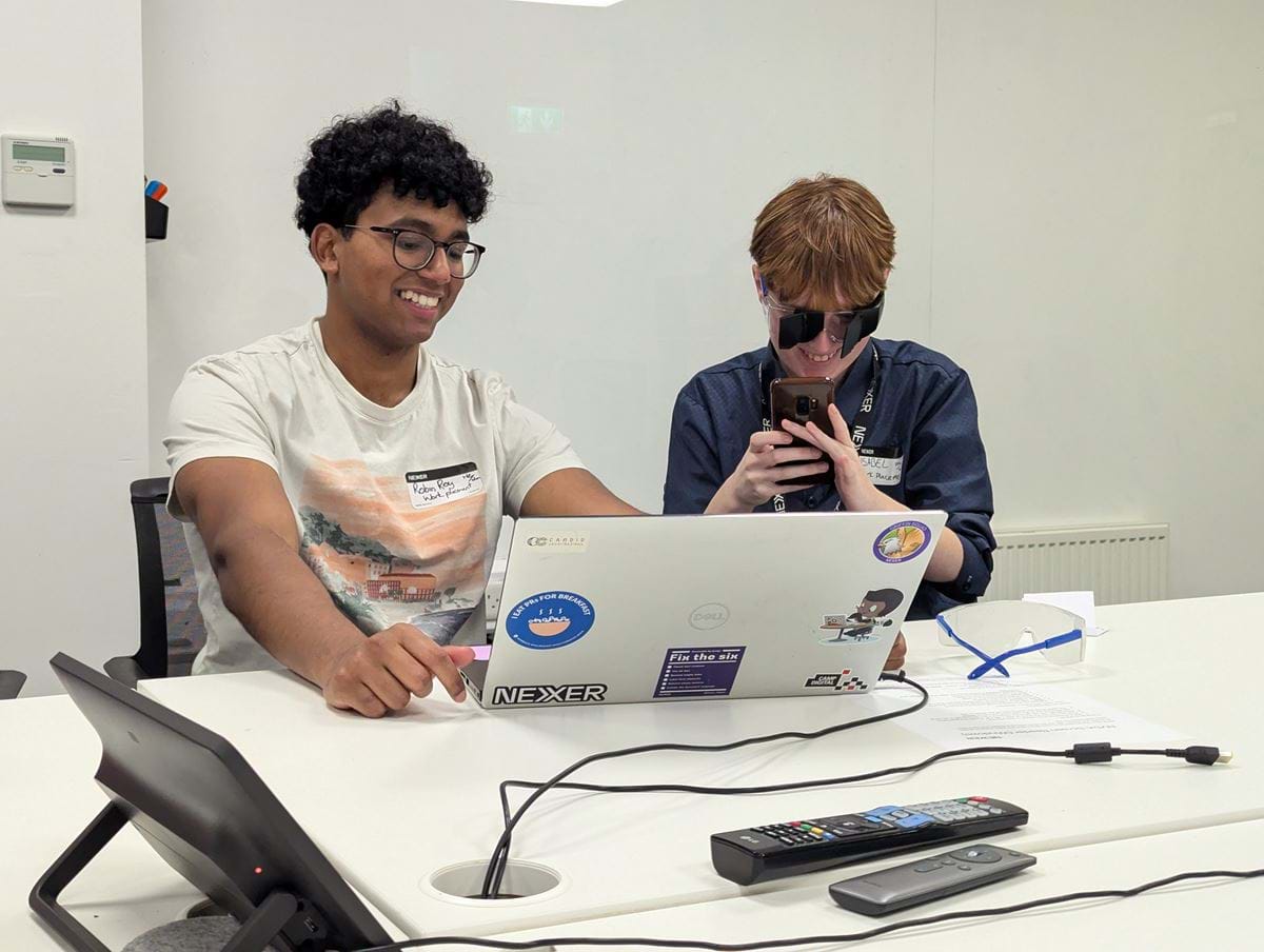 Robin and Isobel using the popup accessibility lab. Robin has brown eyes and hair, and is smiling while using a laptop. Isobel has brown hair, and is wearing simulation goggles. On the table is another pair of simulation goggles.