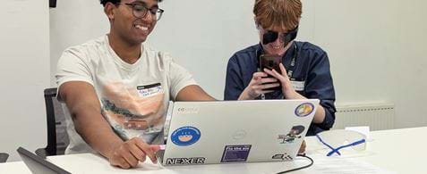 Robin and Isobel using the popup accessibility lab. Robin has brown eyes and hair, and is smiling while using a laptop. Isobel has brown hair, and is wearing simulation goggles. On the table is another pair of simulation goggles.