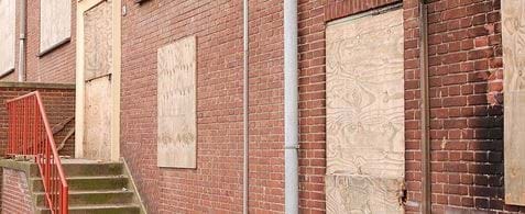 An old red brick building, with stone steps and a red railing at the front. All the windows and doors are boarded up