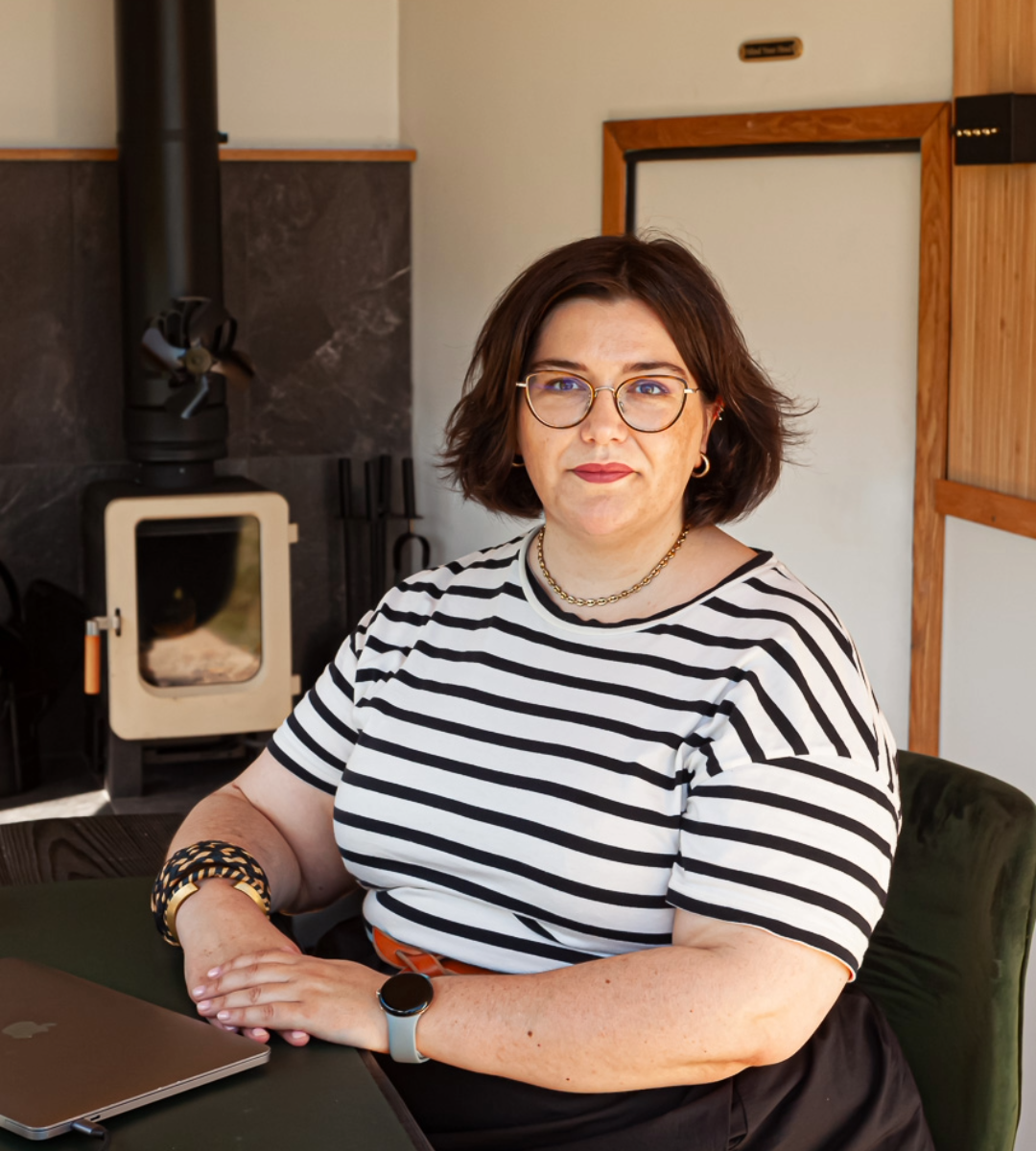 Portrait of a person with short dark hair and glasses, wearing a black-and-white striped top and sitting at a table with a laptop. A wood-burning stove is visible in the background.