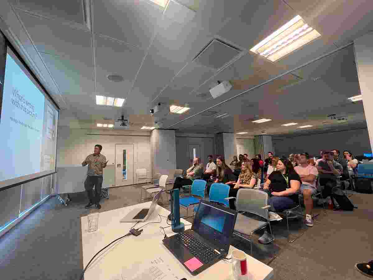 Chris Bush standing in front of a seated audience of people and delivering a presentation. A projected slide reads "What's wrong with this picture?"