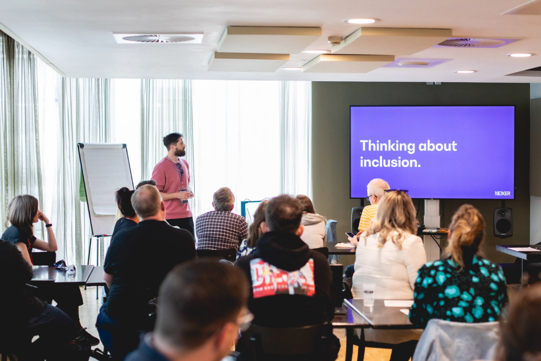 Danny Lancaster stands in front of a room of seated people. Onscreen a slide reads 'Thinking about inclusion'