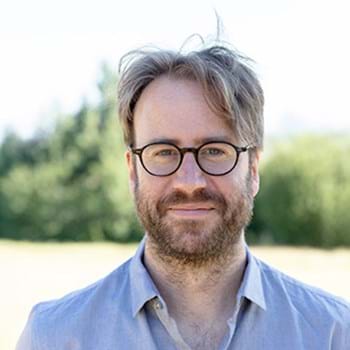 Richard is wearing glasses, has a beard, and stands outdoors, wearing a light blue shirt He is standing in an open area surrounded by trees and a on a sunny day.