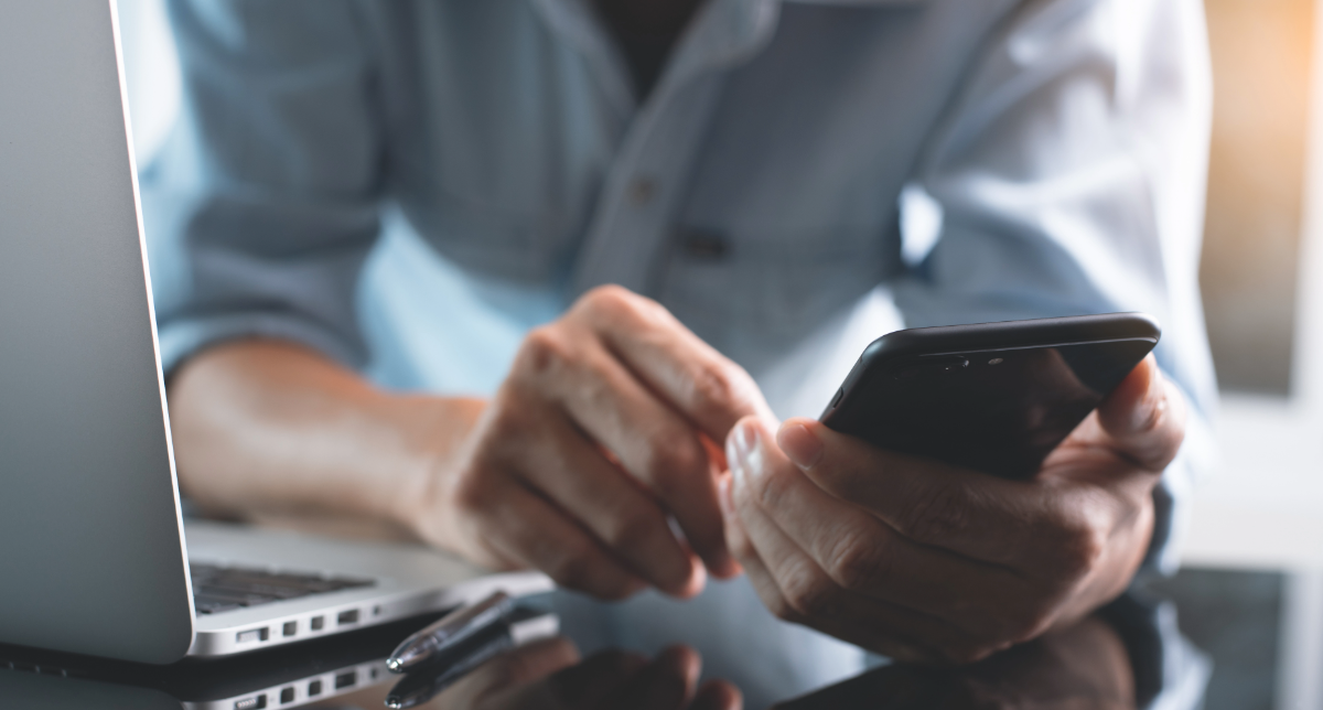 Close‑up of hands using a smartphone next to an open laptop on a desk, representing everyday digital work across multiple devices.