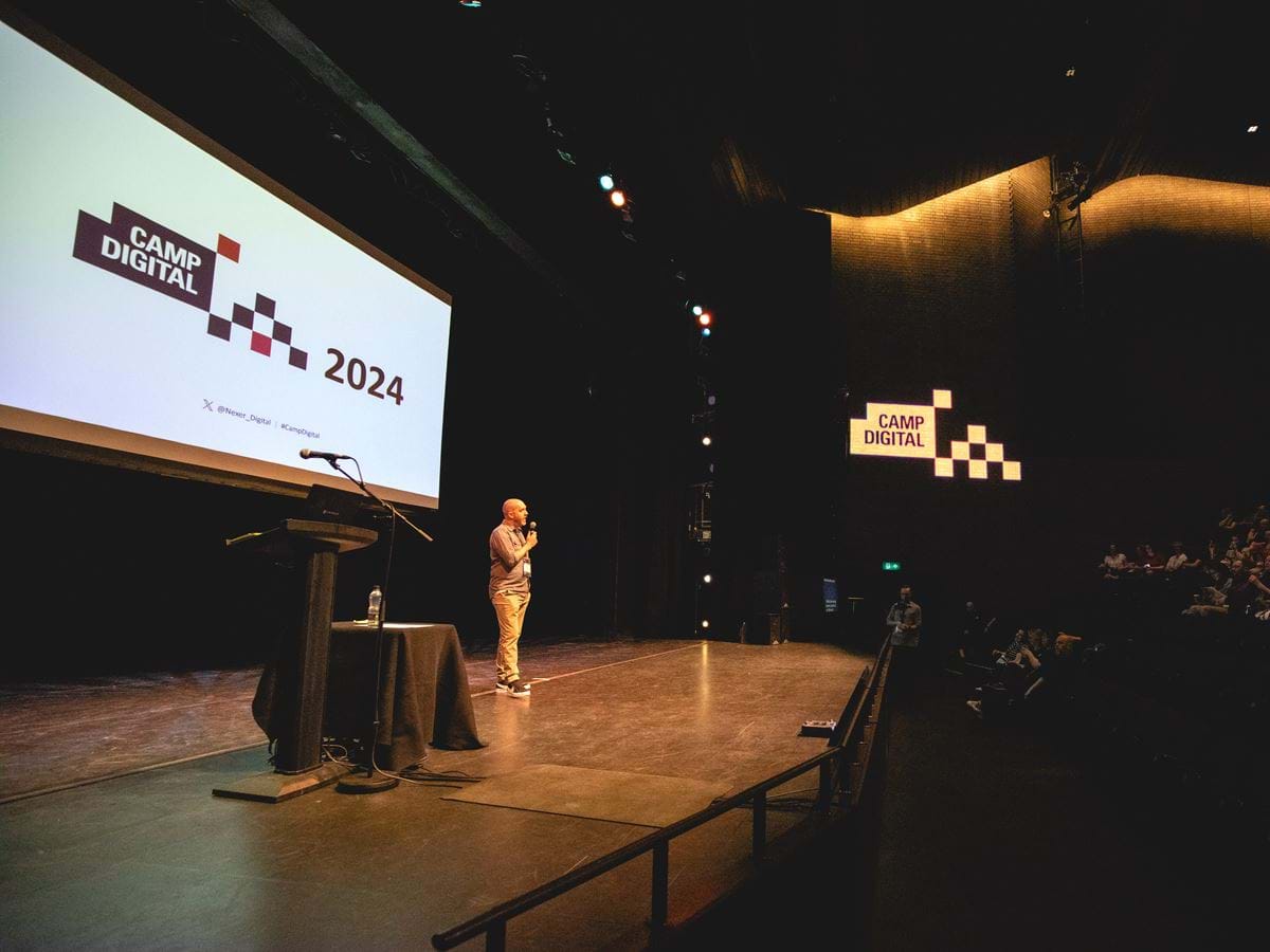 Camp Digital host Shaun Gomm stands onstage at the RNCM, with the Camp Digital logo visible behind and on the wall