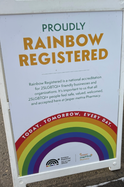Sign reads "Proudly rainbow registered. Rainbow registered is a national accreditation for 2SLGBTQI+ friendly businesses and organisations. It's important to us that 2SLGBTQI+ people feel safe, valued, welcomed, and accepted here at Jasper Mettra Pharmacy.