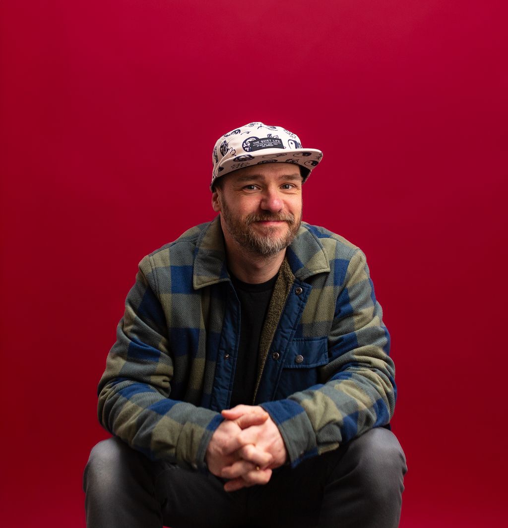 Portrait of Gavin Strange seated against a bright red background, wearing a patterned cap and checked jacket.