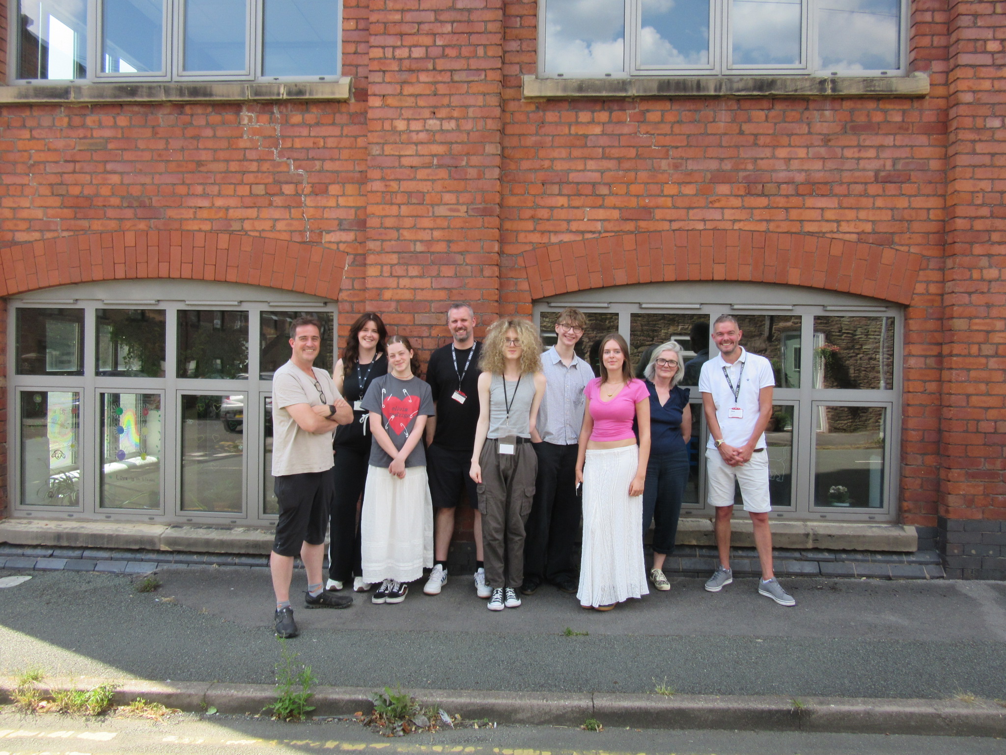 A group of nine people stand outside a red-brick office building, spaced across the entrance and posing for a group photo. The central doorway features a large window with colourful geometric artwork visible behind the glass. Above the entrance is a stone sign reading "James Kershaw". The group includes both Nexer Digital team members and work experience students, dressed in casual clothing, standing on the pavement in front of the building on a bright day.