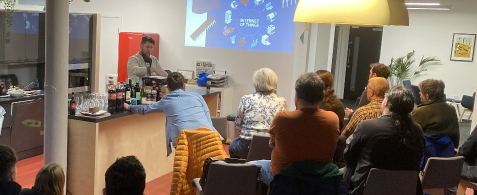 A group of people seated in the Nexer offices. They're watching a person stood presenting a slide.