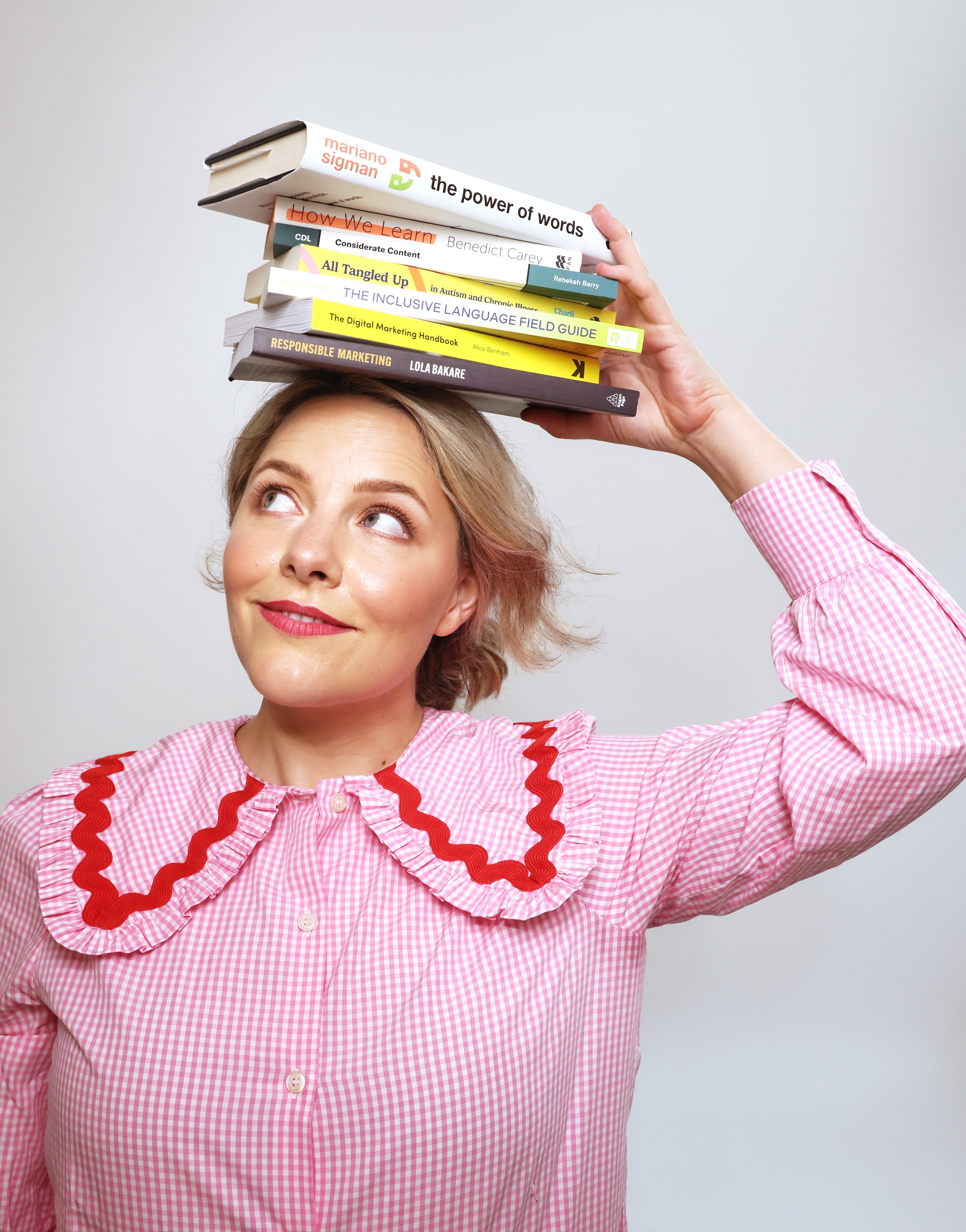 Studio portrait of Ettie Bailey-King wearing a pink top and balancing several books on their head while looking upwards.