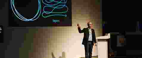 Matt Edgar, a white man dressed in a casual suit and trainers, standing on stage with a presentation screen in the background. There's a squiggly line on the screen and with the word, "Layers" written below it. 