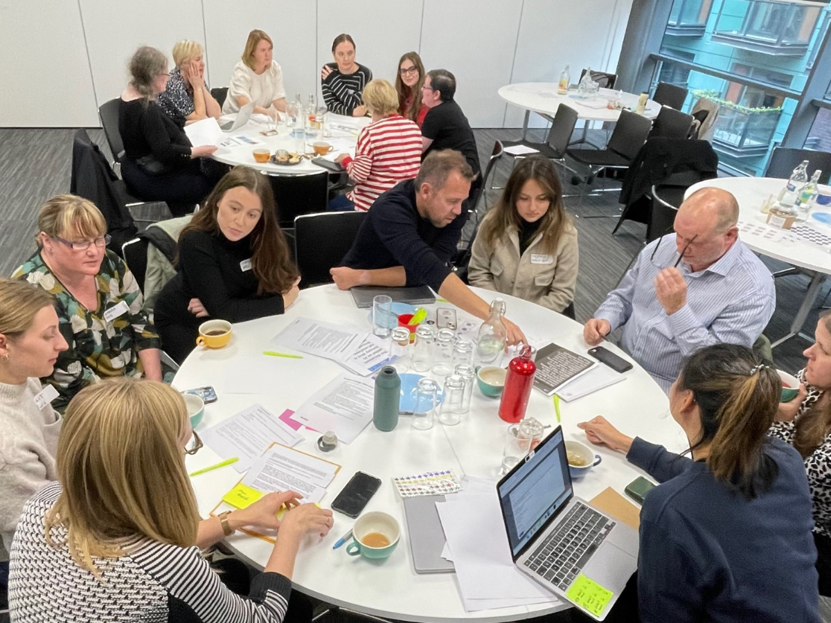 A group of people seated around two circular tables. They are writing on notepads, laptops and post-it notes.