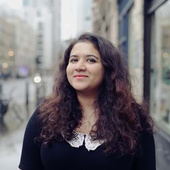 Hera grins at the camera. She has long, curly hair. Stands on a city street, wearing a black top with a white lace collar. The background is blurred, showing buildings and parked bicycles on a rainy day