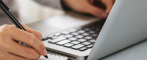 A person writing in a notepad and typing on a laptop