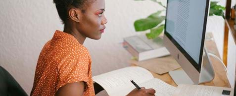 A woman is looking at a laptop screen and making notes in a notepad