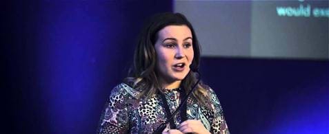 Molly Watt. A white woman with long brown hair, wearing a printed top, speaking into a microphone and standing at a podium. She is stood against a blue background.