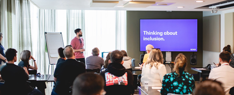 A workshop space at Camp Digital, with accessibility team lead Danny Lancaster addressing a room of seated people. Onscreen is a slide reading "Thinking about inclusion"