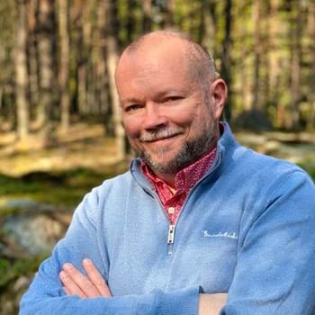 Per is crossing his arms while standing happily in a sunlit forest. He is wearing a light blue sweater over a red patterned shirt. The background features tall trees and greenery