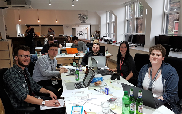 A group of white people sat around a table in an office all looking at the camera, with laptops open in front of them.