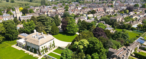 An aerial shot of Blenheim Palace, with the grand green gardens surrounding it. Beyond that houses can be seen stretching into the distance.