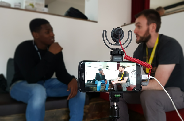 A smartphone in the foreground set up on a tripod, recording one black man and one white man, having a chat, in the background.