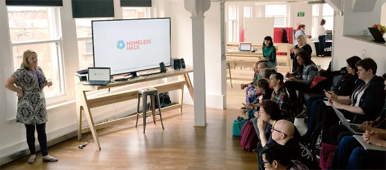 Woman standing next to a large screen giving a presentation to a group of people sitting opposite her