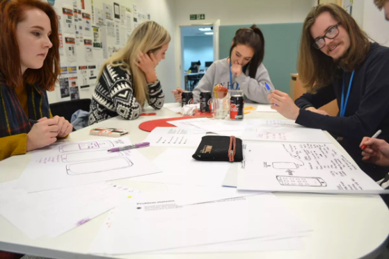 Four people, three women and one man, sitting around a desk working collaboratively and looking at sketches and diagrams