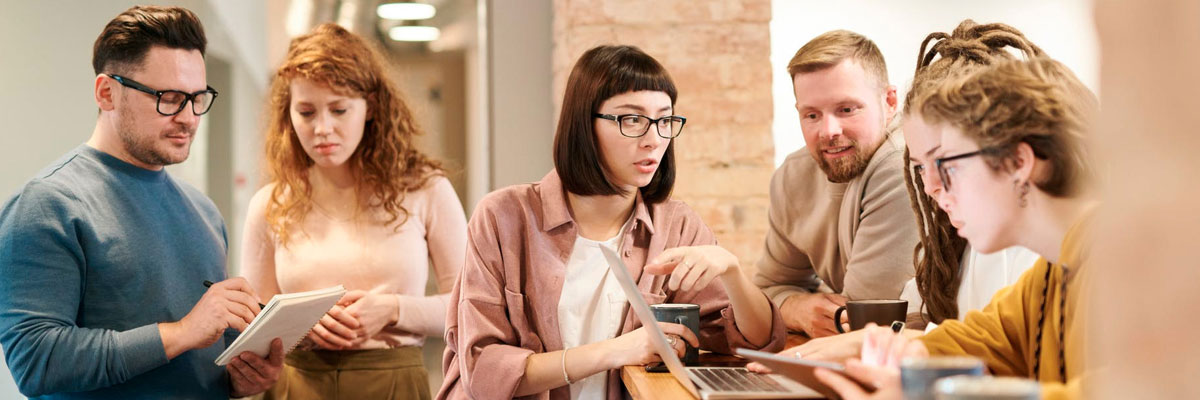 Two groups of people stood working around laptop and a notepad.