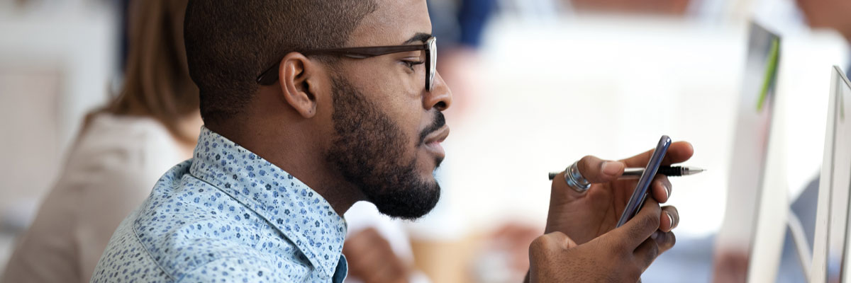 Man in front of computer holding phone in hand and looking at the screen